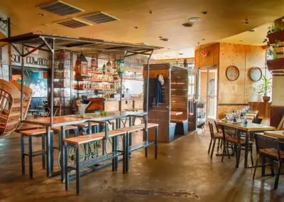Bright, wood‑paneled restaurant interior with long communal tables and stools on the left, a stocked backlit bar and hanging lanterns in the center, and smaller dining tables with place settings near windows on the right.