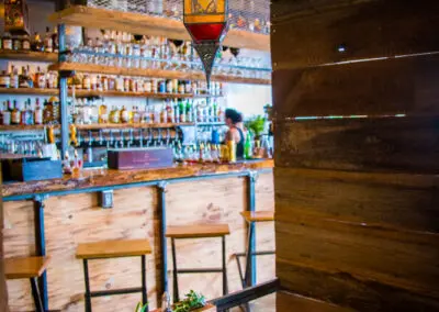 A cozy restaurant booth with a wooden table set for two, patterned cloth napkins and a small condiment bottle on the table, Moroccan-style hanging lantern above, and a wood-and-metal bar with stools and shelves of bottles in soft focus in the background