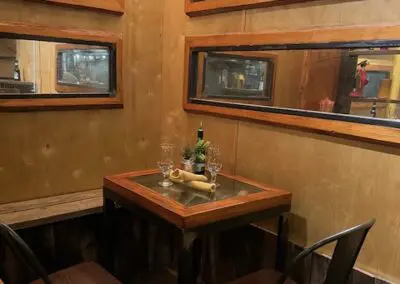 Small rustic dining nook with a square wooden table set for two (wine bottle, two wine glasses, small plant, bread roll) surrounded by wooden bench and two metal chairs, warm string lights and rectangular mirrors on paneled walls reflecting the cozy interior