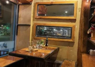 Cozy wooden restaurant corner with a small table set for two—empty wine glasses, a bottle, napkin and cutlery—benches, three horizontal framed windows on a tan wall and string lights above, night street visible through glass.