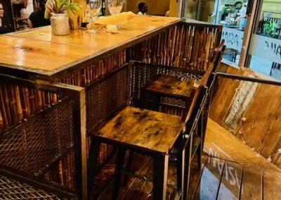 Rustic wooden bar counter with three high stools, place settings with wine glasses and a pineapple centerpiece, warm ambient lighting and a street-facing window showing passersby.
