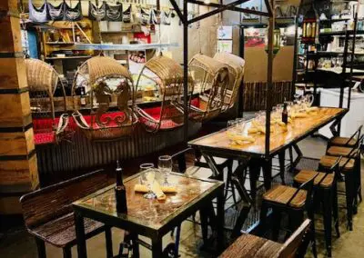 Cozy restaurant interior with wooden communal tables and benches, a bicycle front wheel attached to a table, hanging rattan basket chairs along a corrugated metal wall, warm overhead lighting, place settings with glasses and napkins, and shelves of bottles and condiments in the background