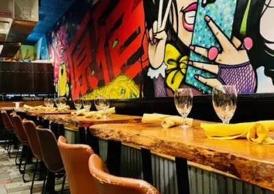 A long wooden bar counter set for dining with folded yellow napkins and wine glasses, a row of brown leather bar stools, tiled floor, and a colorful graffiti mural featuring bold red characters and stylized comic-style artwork along the wall.