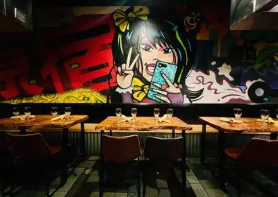 Restaurant interior with three wooden tables set with wine glasses and napkins, row of leather chairs, and a large colorful graffiti mural of a stylized girl making a peace sign on the back wall.