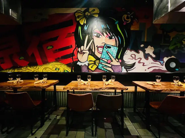 row of leather bar chairs at a live-edge wooden counter set with wine glasses and yellow napkins against a colorful Asian street-art mural wall in an industrial-chic sushi and noodle restaurant interior
