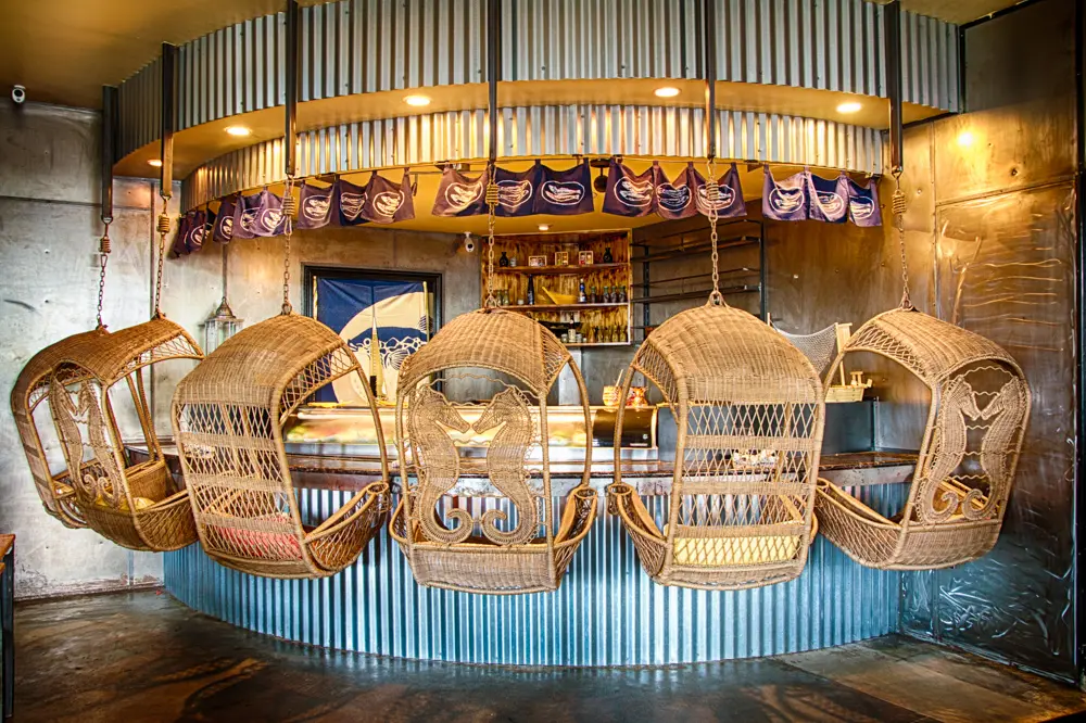 Row of six wicker hanging basket chairs suspended at a corrugated metal bar counter in a warmly lit cafe interior