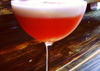 Cocktail coupe filled with a frothy pink-red cocktail garnished with a halved strawberry on the rim, shot on a wooden bar surface for Yaya's Cookbook cocktails collection