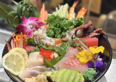 Bowl of colorful sashimi and sushi ingredients on a restaurant counter: assorted raw fish slices (tuna, white fish, salmon), octopus, shrimp, avocado slices, lemon wedges, pickled ginger, wasabi, tobiko roe, shredded carrot, kale garnish, edible flowers and a purple orchid, arranged over rice in a black bowl for a seafood dinner recipe.