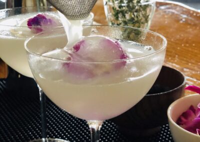 Cocktail being strained into a coupe glass over a large round ice sphere with a purple edible flower, another identical cocktail behind it, a bartender’s metal strainer at top left, and a small bowl of flower petals on a bar mat with bright window light and a vase of herbs in the background